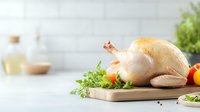 Raw whole chicken on wooden cutting board with fresh herbs and vegetables against light kitchen background, ready for cooking preparation.
