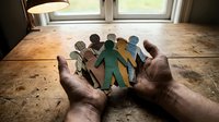 Hands holding diverse paper cutout figures representing unity, teamwork and community support in warm natural lighting on wooden surface.