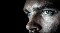 Close up portrait of young Caucasian male with striking bright blue eyes in dramatic dark lighting, showing partial face profile against black background.