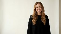 Young Caucasian woman with long wavy brown hair wearing black sweater smiling at camera against neutral beige wall background in modern interior space.