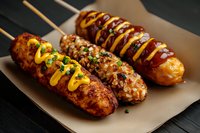 Grilled corn dogs and sausages on wooden sticks topped with mustard sauce, chopped herbs and spices served on brown craft paper. Close up food photography.