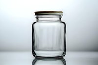 Empty glass jar with metal screw lid on reflective surface against light background, minimalist composition for food storage and preservation concepts.