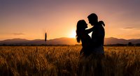 Romantic couple silhouette embracing in golden wheat field at sunset with mountains and tower in background creating intimate moment for love concepts.