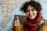 Young Mediterranean woman with curly hair wearing red knitted scarf and yellow sweater showing thumbs up against world map background, expressing positivity and approval.
