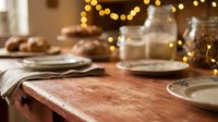 Wooden dining table set for holiday celebration with warm golden string lights, plates, glasses, and festive decorations in soft focus background for seasonal entertaining.