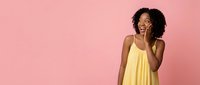 Young African American woman in yellow dress covering mouth with hand in surprise gesture against pink background for advertising and lifestyle content.