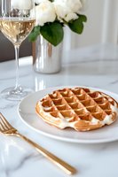 Golden Belgian waffle on white plate with glass of wine and gold cutlery on marble surface, white roses in silver vase create elegant breakfast setting.
