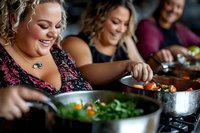 Curvy woman enjoying cooking fresh vegetables with friends in kitchen, sharing healthy meal preparation and social dining experience with genuine smiles.