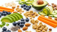 Healthy organic food ingredients with avocado, blueberries, nuts, seeds and vegetables arranged on white background for nutritious meal planning.