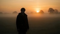 Young man standing alone in misty field at golden sunset, dramatic silhouette against glowing orange sky with fog and bare trees.