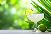 Refreshing daiquiri cocktail with lime garnish and palm leaves on wooden surface against blurred green tropical background, summer drink styling.