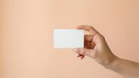 Close-up of a hand holding a blank white business card against a neutral beige background. Suitable for branding, corporate identity, and marketing materials.