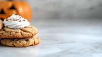 Homemade pumpkin spice cookies topped with whipped cream and sprinkles on marble surface, with decorative orange pumpkin in background for autumn baking.