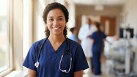 Young African American female nurse in blue scrubs with stethoscope smiling confidently in hospital corridor with medical staff in background.
