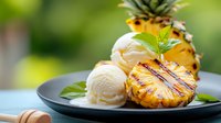 Grilled pineapple slices served with vanilla ice cream scoops and mint garnish on black plate against blurred green background, summer dessert.