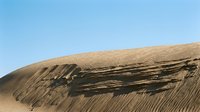 Massive sand dune with dramatic wind-carved ridges and striations under clear blue sky, showcasing desert landscape erosion patterns and natural geological formations.