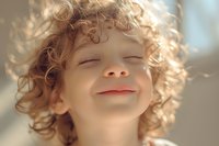 Young child with curly blonde hair smiling with eyes closed in warm sunlight, expressing pure joy and innocence. Natural lifestyle portrait capturing genuine happiness.