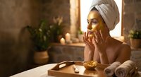 Young woman with towel on head applying face mask in spa bathroom with candles and plants for skincare routine and wellness relaxation.