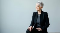 Senior woman with short gray hair wearing black blazer and dark top, sitting against neutral background, professional business portrait for corporate and executive use.