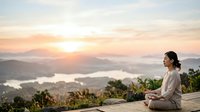 Woman in casual white shirt meditating on mountain overlook at sunrise, gazing at misty valley landscape with lakes and forested hills below.