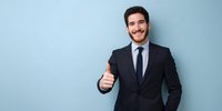 Confident young businessman in formal suit showing thumbs up gesture against light blue background, expressing approval and success.