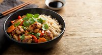 Asian stir-fry with chicken, vegetables and rice in black bowl on wooden table with chopsticks and soy sauce for healthy dining and cooking concepts.