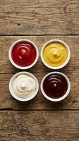 Four different condiment sauces in white bowls on rustic wooden table background for food photography and culinary design projects.