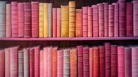 Row of vintage hardcover books arranged on wooden shelves in shades of pink, coral, and purple creating an aesthetic color-coordinated library display. Decorative spines form pattern.