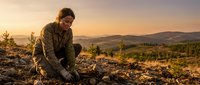 Male farmer or agricultural worker in brown jacket planting seeds in fertile soil at sunrise, with rolling hills and forest landscape in background, suitable for farming, agriculture, and sustainability content.