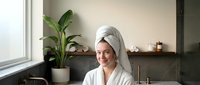 Young Caucasian woman in white bathrobe and towel on head smiling in modern bathroom after shower, spa self-care routine.