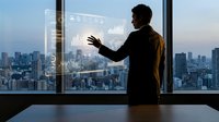 Professional businessman interacting with futuristic holographic interface overlooking modern city skyline through office window at sunset.