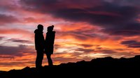 Silhouetted couple standing together against dramatic orange and red sunset sky with mountain landscape, romantic evening scene for relationship and love concepts.
