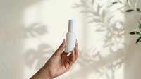 Unisex hand holding white cosmetic spray bottle against neutral background with natural plant shadow overlay for skincare or beauty product mockup.