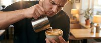 Middle-aged male barista with light skin tone pouring steamed milk into espresso cup, creating latte art in warm cafe setting with blurred background and ambient lighting.