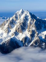 Majestic snow-covered mountain peak rising above clouds, dramatic rocky cliffs and ridges illuminated by sunlight against blue sky, winter alpine landscape.