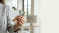 Woman in business attire holding blank white card in modern office environment for corporate messaging, branding, or promotional content display.