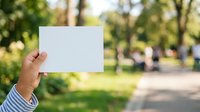 Person holding blank white card in park with green trees and blurred background, suitable for advertising, marketing mockups, promotional campaigns, and outdoor business presentations.