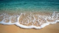 Gentle ocean wave rolling onto sandy beach with white foam creating natural lace pattern against turquoise blue water. Perfect for travel and relaxation themes.