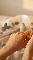 Woman applying golden facial serum oil with dropper for skincare routine and anti-aging treatment in warm natural light setting.