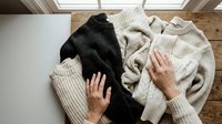 Hands organizing cozy knit sweaters in neutral tones on wooden floor. Seasonal wardrobe preparation and clothing storage concept for home organization.