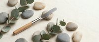 Wooden handle palette knife with eucalyptus leaves and smooth river stones arranged on neutral textured background for art supplies and natural decor.
