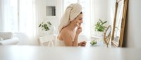Young woman with towel on head applying skincare routine in bright modern bathroom mirror reflection for wellness and self-care morning ritual.