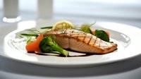 Grilled salmon fillet served with steamed broccoli, baby carrots and lemon slice on white plate, garnished with fresh herbs against light background.