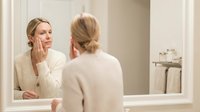 Middle-aged Caucasian woman examining her face in bathroom mirror wearing cream sweater for skincare routine and anti-aging beauty concept.