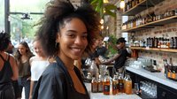 Young African American woman with curly hair smiling at trendy bar with rustic brick walls and shelves of bottles, capturing social atmosphere in restaurant setting.