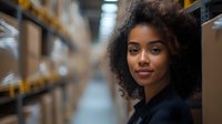 Young African American female professional with natural curly hair in warehouse logistics environment looking confidently at camera, selective focus background.