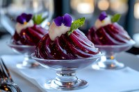 Elegant dessert featuring sliced red beets layered in crystal glass, topped with cream and purple edible flowers, served on white tablecloth in upscale setting.
