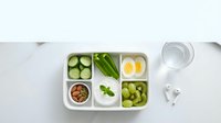 Healthy green lunch box with compartments containing fresh vegetables, boiled eggs, nuts and water for balanced nutrition and meal prep.