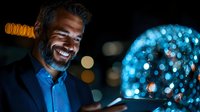 Mature businessman with gray beard smiling while looking at glowing blue sphere with digital network visualization against dark background with bokeh lights.