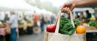 Shopper holding canvas bag filled with fresh produce at farmers market with blurred crowd and vendor stalls in background for retail and agriculture marketing.